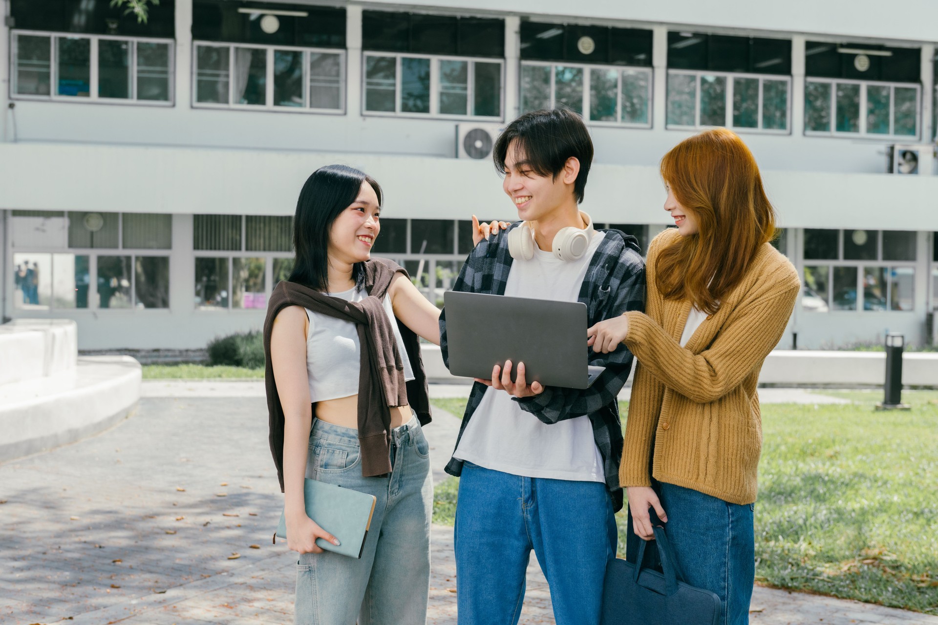 Three cheerful young adult students are discussing a project on a laptop outside their university, enjoying the pleasant weather and collaborative learning environment